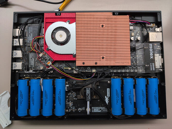 The mnt reform laptop from the back, with the transparent bottom plate removed. Next to the large heat sind, a fan has been mounted using a 3D printed bracket. A black PCB has also been mounted to the same bracket. It is connected to both the fan and also the motherboard via colorful cables.